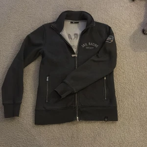 Svart full zip tröja från Sail Racing - Säljer en grå full zip tröja från Sail Racing med dragkedja och broderad logga på bröstet och ryggen. Jackan har långa ärmar och ribbade muddar. Perfekt för en stilren look.