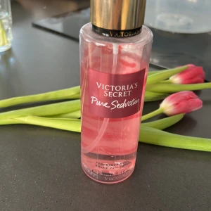 Victoria's Secret Pure Seduction Fragrance Mist - Säljer en Victoria's Secret Pure Seduction Fragrance Mist med en doft av saftig plommon och krossad fresia. Flaskan är genomskinlig med rosa vätska och har en gyllene kork. Perfekt för att fräscha upp din dag med en söt och blommig doft.