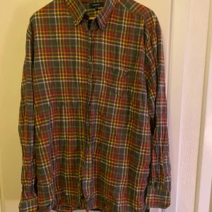 Rutig skjorta från GANT - Säljer en färgglad rutig skjorta från GANT i storlek 2XL. Skjortan har långa ärmar och knappar framtill. Perfekt för en avslappnad stil med sina jordnära färger i grönt, rött och orange.