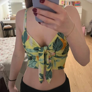 Grön blommig knyt topp - Säljer en somrig grön blommig topp med knyt framtill. Toppen har smala axelband och en croppad passform, perfekt för varma dagar. Den har ett färgglatt mönster med inslag av gult och orange.💚