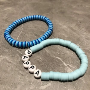 Två armband med bokstavspärlor - Säljer två fina armband. Ett i blå och ljusblå randigt mönster och ett ljusblått med vita bokstavspärlor som bildar ordet 'PAPPA'. Perfekt som en personlig present eller för att bära själv. 100kr för båda 50kr för ett 