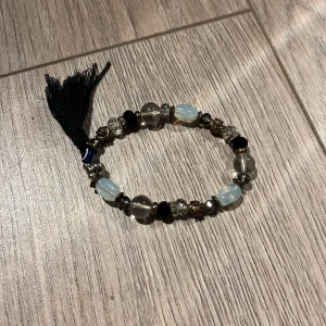 Armband med pärlor och tofs - Unikt armband med en mix av pärlor i olika färger och material, inklusive genomskinliga och svarta pärlor. Armbandet har en dekorativ svart tofs som ger en bohemisk touch. Perfekt accessoar för att lyfta din stil.