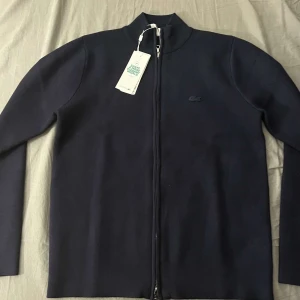 Svart tröja med dragkedja från Lacoste - Säljer en stilren svart tröja från Lacoste med dragkedja framtill. Tröjan har långa ärmar och en diskret logga på bröstet. Perfekt för en avslappnad och snygg look.