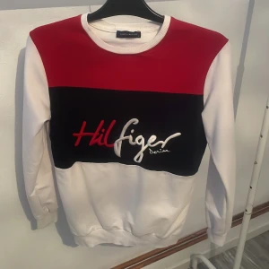 Vit och röd tröja Tommy Hilfiger - Säljer en snygg vit och röd tröja från Tommy Hilfiger med svart panel och broderad logga på framsidan. Tröjan har långa ärmar och en bekväm passform. Perfekt för en stilren look.