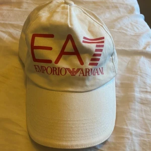 Beige keps från Emporio Armani - Snygg beige keps från Emporio Armani med röd EA7-logga framtill och 'ITALIA' broderat i grönt baktill. Justerbar rem med italienska flaggans färger för perfekt passform.