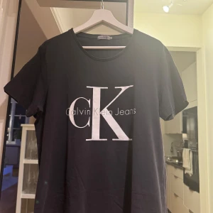 Svart t-shirt från Calvin Klein Jeans - Snygg svart t-shirt från Calvin Klein Jeans med stort vitt CK-logotryck på framsidan. Perfekt för en stilren och avslappnad look. Gjord i mjukt material för extra komfort. Passar till allt. Skick 7,5/10,storlek xl sitter som liten L!