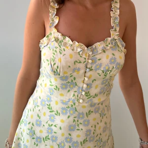 Blommig sommarklänning  - Söt sommarklänning med blommigt mönster i ljusgult och blått. Klänningen har volangdetaljer vid axelbanden och knappar framtill. Perfekt för soliga dagar!