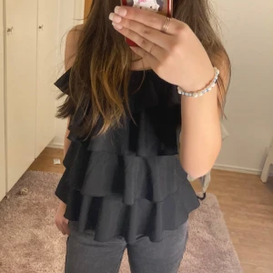 Svart volangtopp - Snygg svart topp med flera lager volanger. Passar mig som har storlek S. Perfekt för en stilren look. Toppen har smala axelband och en luftig passform.