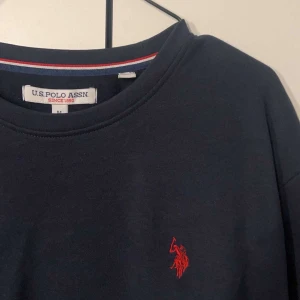 Mörkblå tröja från U.S. Polo Assn. - Säljer en stilren mörkblå tröja från U.S. Polo Assn. med ett rött broderat emblem på bröstet. Tröjan har långa ärmar och en rund halsringning. Perfekt för en avslappnad look. Använt en gång.