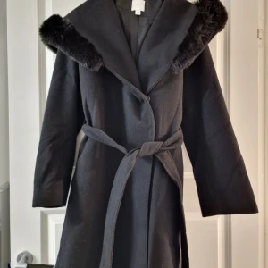 Svart kappa med pälsdetaljer - En helt oanvänd svart kappa med mjuk pälsdetalj runt kragen. Köpt på HM några säsonger sedan för 900 kr. Kappan har ett knytbälte i midjan och knappar framtill. Perfekt för kyliga dagar🤗 passar mig som har s/m