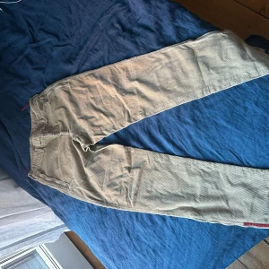 Beige flare manchesterbyxor från Prada - Snygga beige manchesterbyxor från Prada med en stilren design. Byxorna har en dragkedja vid bensluten och en diskret logga i rött. Perfekta för en trendig flared look. 