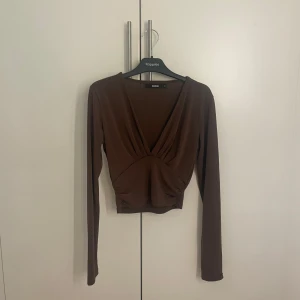 Brun långärmad topp - Säljer en stilren brun långärmad topp från Bikbok med v-ringad design. Perfekt för en chic och enkel look. Toppen är i ett mjukt och bekvämt material.