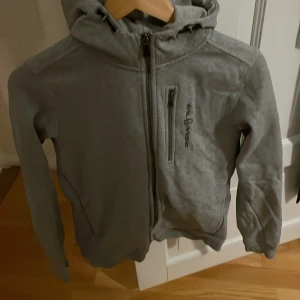 Grå huvjacka från Sail Racing - Säljer en grå huvjacka från Sail Racing med dragkedja framtill och en praktisk bröstficka med dragkedja. Jackan har en broderad logga på bröstet och en på huvan. Perfekt för en casual look.