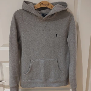 Grå hoodie Ralph Lauren stl 14-16 - Säljer en klassisk hoodie från Ralph Lauren, gråmelerad med marinblå logga. Stl 14-16, se mått. Superskick, knappt använd, tyget känns helt nytt! Mjukaste fleecekänsla på insidan. Mått: Armhåla till armhåla 48cm, bredd längst ner 46cm, längd från mitt på axeln 57,5cm.