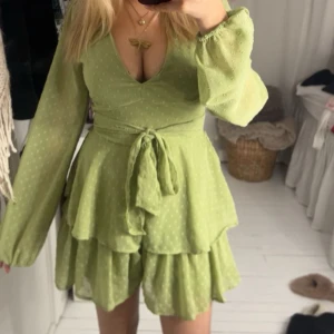 Grön klänning  - Säljer en söt grön klänning från New Yorker med v-ringning och långa ärmar. Klänningen har ett knytband i midjan och volangdetaljer som ger en lekfull look. Även inbyggda shorts.