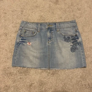 Jeanskjol med broderi - Snygg jeanskjol i ljusblå denim med broderade blommor på fram- och baksida. Kjolen har en rå kant nertill och slitna detaljer.