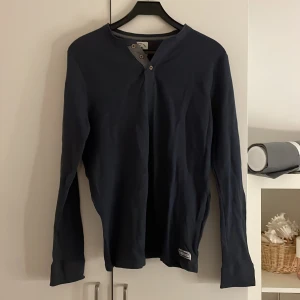 Mörkblå långärmad tröja från Denim Factory - Säljer en stilren mörkblå långärmad tröja från Denim Factory. Tröjan har en knappdetalj vid halsen och en liten etikett nertill. Perfekt för en avslappnad look.