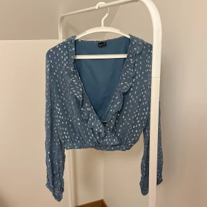 Blå omlottblus från Gina Tricot - Säljer en blå omlottblus från Gina Tricot med vita prickar och volangdetaljer. Blusen har långa ärmar och en v-ringad design. Perfekt för en stilren look.