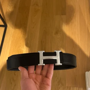 Svart skärp med silverfärgat H-spänne - Snyggt svart skärp i skinn med ett stilrent silverfärgat H-format spänne. Perfekt för att ge en elegant touch till din outfit. Skärpet har en klassisk design och passar till många olika stilar.