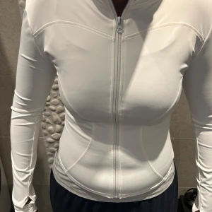 Liknade lululemon tröja - Säljer en vit långärmad träningströja med dragkedja framtill. Tröjan har en tight passform och är tillverkad i ett syntetmaterial som andas. Perfekt för träning och andra aktiviteter. Den har även hål för tummarna vid ärmsluten för extra komfort.