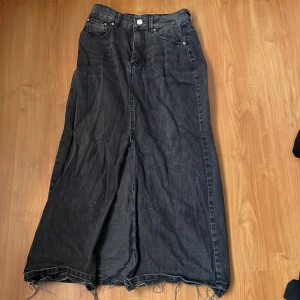 Svart jeanskjol - Snygg svart långkjol i jeansmaterial med rå kant. Kjolen har en klassisk femficksdesign och en knapp framtill. Perfekt för en avslappnad stil.