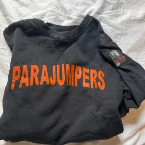 Svart fleece tröja från Parajumpers - Säljer en svart fleece tröja från Parajumpers med orange text på framsidan. Tröjan har långa ärmar och ett märke på ärmen. Perfekt för alla dagar.