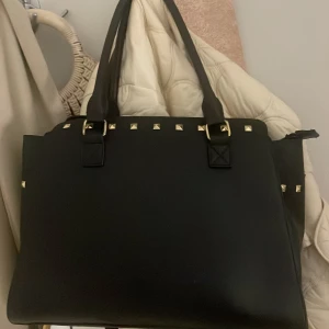 Svart axelväska med nitar - Snygg svart axelväska med guldfärgade nitar som detaljer. Väskan har dubbla handtag och en dragkedja på baksidan för extra förvaring.