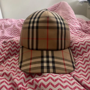 Äkta keps från Burberry - Snygg keps från Burberry i klassiskt mönster. Kepsen har en justerbar rem baktill och är märkt med 'Burberry London England'. Perfekt för en stilren look. Knappt använd och köpt för 4500