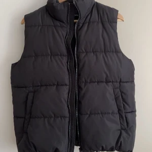 Svart dunväst med dragkedja - Snygg svart dunväst med dragkedja framtill. Perfekt för kyligare dagar och enkel att matcha med olika outfits. Västen har en hög krage och två sidofickor för extra bekvämlighet. (EGNA BILDER PRIVAT) 