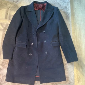 Mörkblå  kappa med dubbelknäppning - Elegant mörkblå kappa med dubbelknäppning och breda slag. Kappan har en stilren design med knappar både fram och bak samt långa ärmar. .