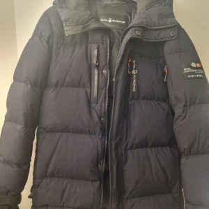 Svart dunjacka från Sail Racing - Svart dunjacka från Sail Racing med broderad logga och detaljer på ärmen. Jackan har en hög krage och dragkedja framtill. 