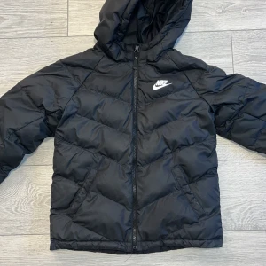 Svart dunjacka från Nike - Säljer en svart dunjacka från Nike med huva och dragkedja. Jackan har en klassisk pufferdesign och är perfekt för kyligare väder. Den är märkt med Nike-loggan på bröstet och har en bekväm passform. På sista bilden kan man se slitage på jackan, vill ni ha fler bilder så hör av er. Köptes vintern år 2022🙂