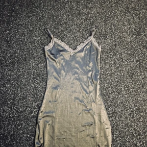 Grå satin klänning  - Snygg grå satin klänning från Divided med spetsdetaljer vid halsringningen. Klänningen har smala axelband och en figurnära passform. Perfekt för en stilren look. Sitter skönt mot kroppen.