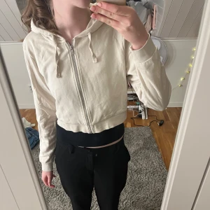 Vit croppad hoodie - Säljer en vit croppad hoodie med dragkedja och huva. Den har långa ärmar och ribbade muddar. Perfekt för en avslappnad stil.