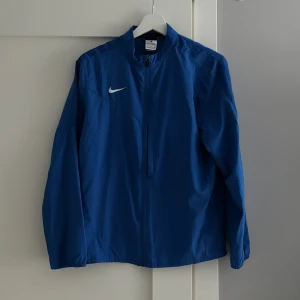 Blå vindjacka från Nike - Säljer en stilren blå vindjacka från Nike med dragkedja framtill och en liten vit logga på bröstet. Jackan har långa ärmar och en hög krage, perfekt för blåsiga dagar. Passar bra för sportiga aktiviteter eller som en lätt jacka till vardags. Hör av er vid intresse!