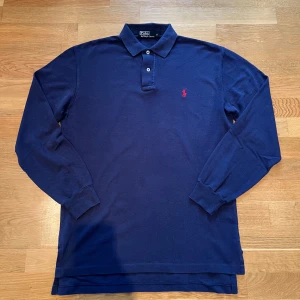 Mörkblå pikétröja från Ralph Lauren - Säljer en klassisk mörkblå pikétröja från Ralph Lauren med den ikoniska röda loggan på bröstet. Tröjan har långa ärmar och knappar vid kragen. Perfekt för en stilren look. Stor i storleken så passar M möjligtvis liten L.