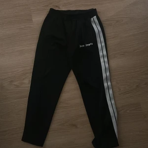 Svarta byxor från Palm Angels - Snygga svarta byxor från Palm Angels med vita ränder längs sidorna. Byxorna har en elastisk midja och är tillverkade i polyester. Perfekta för en avslappnad stil med en touch av lyx.