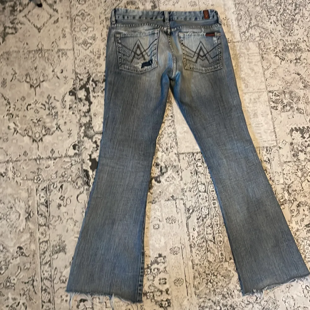 Snygga blå bootcut jeans med slitningar på ena benet och coola detaljer på bakfickorna. Perfekta för en avslappnad stil med en touch av edge. Märket 7 for All Mankind garanterar kvalitet och stil.. Farkut & Housut.