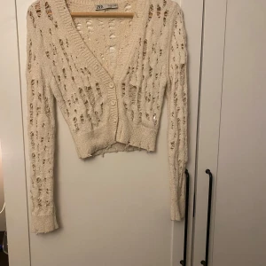 Beige kofta från Zara - Säljer en beige kofta från Zara med ett  hålmönster. Koftan har en v-ringad design och knappar framtill. 