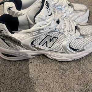 Vita sneakers från New Balance - Snygga vita sneakers från New Balance med silverdetaljer och svart insida.  Aldrig använda. Pris kan diskuteras vid snabb köp