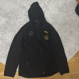 MFF tröja 2021 guld Rakips - Säljer en svart huvtröja från Puma med gulddetaljer. Tröjan har dragkedja och huva, samt logotyper från MFF och Volkswagen på bröstet. Perfekt för sportiga tillfällen eller som en stilren vardagsjacka.