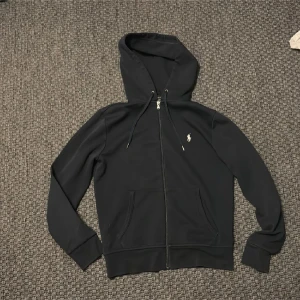 Svart huvtröja från Ralph Lauren - Snygg svart huvtröja från Ralph Lauren med dragkedja och broderad logga på bröstet. Tröjan har långa ärmar och en praktisk huva med dragsko. Perfekt för en avslappnad stil.