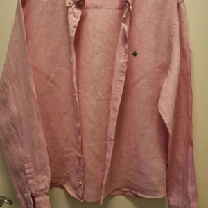 Rosa linneskjorta med broderad detalj - Snygg rosa skjorta i linne från Morris med en liten grön broderad detalj på bröstet. Skjortan har långa ärmar och knappar framtill. Perfekt för en stilren look!