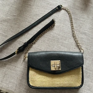 DKNY Svart och beige axelväska med kedja - Snygg DKNY  axelväska i svart och beige med en stilren design. Väskan har en guldfärgad låsdetalj och en kedjerem kombinerad med justerbar axelrem i svart. Perfekt för att ge en touch av elegans till din outfit. Några mindre småskador (se bilder) men inget som stör, därav priset 