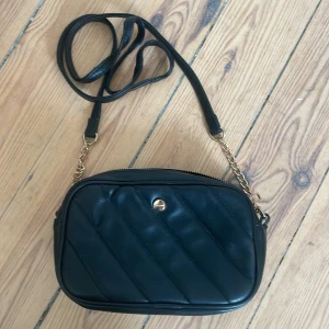Svart väska - Snygg svart axelväska med quiltad design och gulddetaljer. Väskan har en axelrem med kedja och dragkedja upptill. Perfekt för att bära dina nödvändigheter med stil. Har ett extra fak inne i väskan. Använd 1-3 gånger