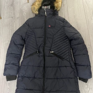 Svart dunjacka med pälskrage - Säljer en svart lång dunjacka från Geographical Norway med pälskrage och dragkedja. Jackan har en quiltad design och långa ärmar. Perfekt för kalla vinterdagar. 🖤