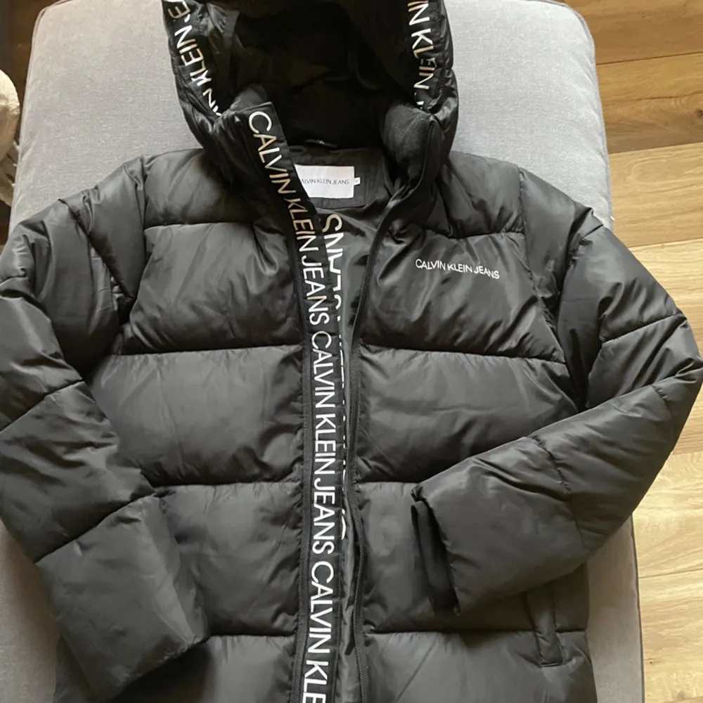 Snygg svart pufferjacka från Calvin Klein med huva och dragkedja. Jackan har märkets logga längs dragkedjan och på bröstet. Perfekt nu under vinter/våren. Storlek 14 år, skulle säga att det motsvarar 164-170. Takit.