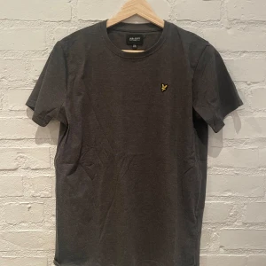 Grå t-shirt från Lyle & Scott - Säljer en stilren grå t-shirt från Lyle & Scott Junior. Den har en liten broderad logotyp i gult på bröstet. Perfekt för en avslappnad look.