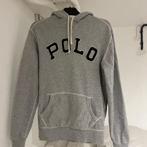 Grå hoodie från Polo Ralph Lauren - Säljer en grå hoodie från Polo Ralph Lauren med stor POLO-text på framsidan. Tröjan har en klassisk känguruficka och justerbar huva med snören. Perfekt för en avslappnad stil.