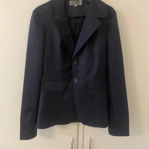 Mörkblå kavaj med knappar - Elegant mörkblå kavaj med två knappar framtill och klassisk krage. Perfekt för en stilren look. Kavajen har långa ärmar och fickor framtill.